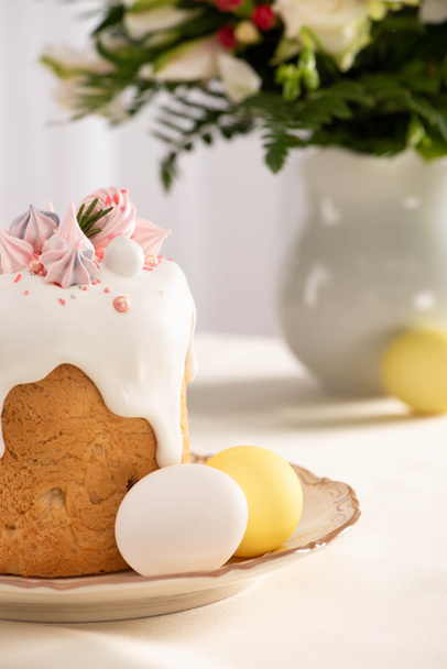 selective focus of delicious Easter cake decorated with meringue near colorful eggs on plate on table with vase of flowers - Photo, Image