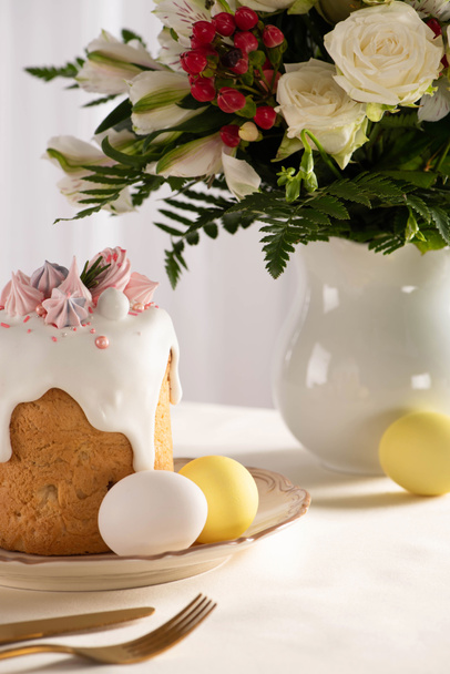 delicioso pastel de Pascua decorado con merengue cerca de huevos de colores en el plato en la mesa con jarrón de flores y cubiertos
 - Foto, Imagen