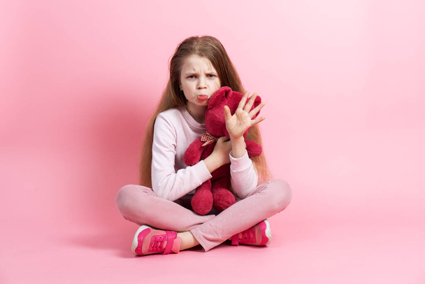 The girl holds a teddy bear in her hands, is naughty and waves her hand. Studio portrait on a pink background. - Foto, Imagem