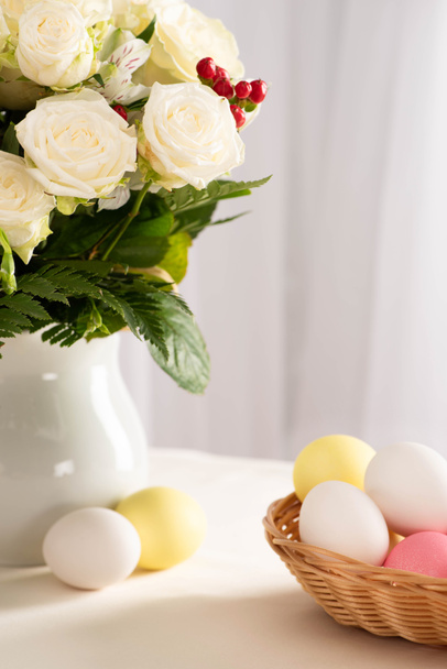 バスケットのカラフルなイースターエッグの近くのテーブルの上に新鮮な花の花瓶 - 写真・画像
