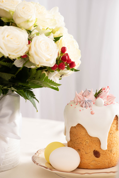 festive Easter cake decorated with meringue near painted eggs on plate and vase of flowers - Фото, изображение