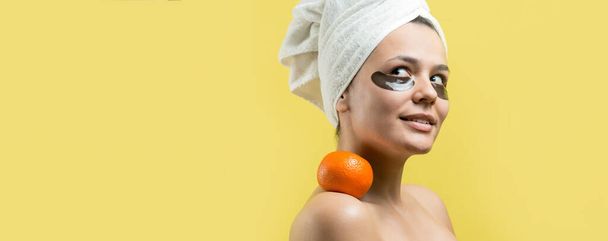 Beauty portrait of woman in white towel on head with gold nourishing mask on face. Skincare cleansing eco organic cosmetic spa relax concept. A girl stands with her back holding an orange mandarin. - Foto, Bild