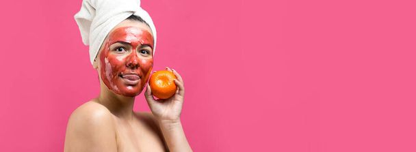 Beauty portrait of woman in white towel on head with red nourishing mask on face. Skincare cleansing eco organic cosmetic spa relax concept. A girl stands with her back holding an orange mandarin. - Photo, image