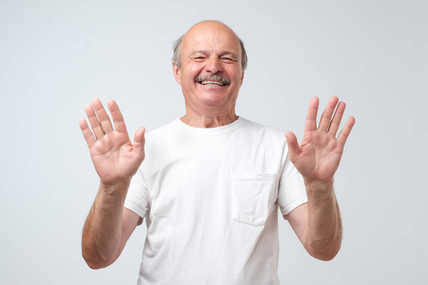 Heyecanlı kel adam beyaz bir arka planda neşeli bir şekilde izole edilmiş on parmağını ve gülüşünü gösteriyor, ellerini kaldırıp onu güldürmeyi kesmesini diliyor. Stüdyo çekimi - Fotoğraf, Görsel