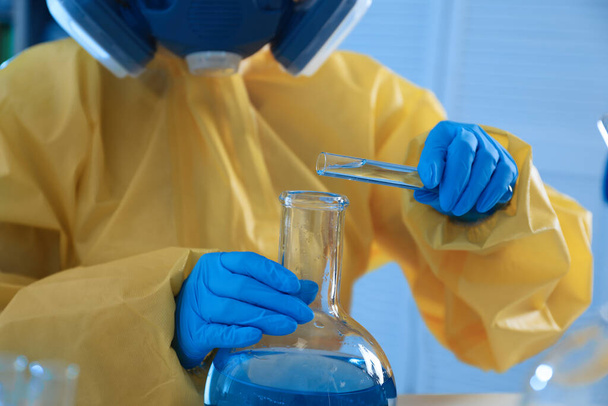 Scientist in chemical protective suit pouring reagent into flask at laboratory, closeup. Virus research - 写真・画像