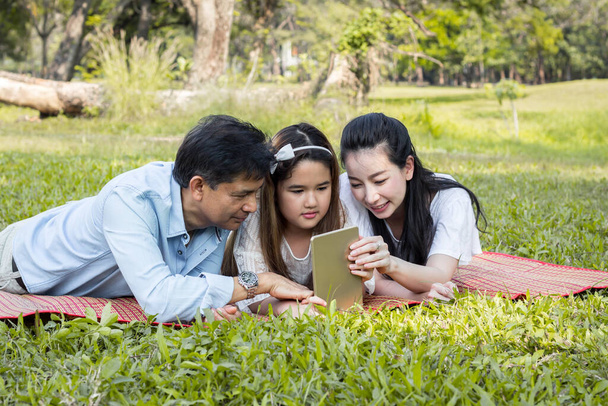 Parents and children are playing the tablet on the mat in the park. Father and mother are teaching by using the tablet. Girls are learning from a tablet that parents are teaching. - Photo, Image