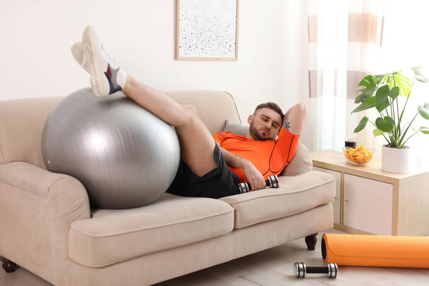 Lazy young man with sport equipment on sofa at home - Foto, Bild
