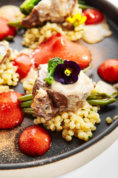 Calf cheeks with ptitim closeup view. Tasty meat with Israeli couscous, sauce and broccoli. Delicious dish decorated with greenery and violets. Restaurant dinner, served food composition - Фото, изображение