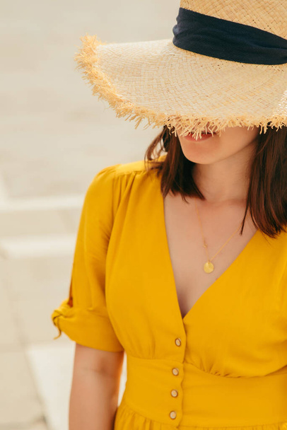 žena ve žlutých slunečních šatech venku skrývá oči za velkou slámou klobouk módní koncept - Fotografie, Obrázek