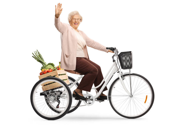 Elderly lady riding a tricycle with a crate full of fruits and vegetables and waving at the camera isolated on white background - Фото, изображение