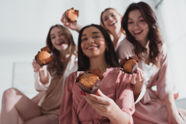 A multikulturális nők szelektív fókusza muffint mutat, mosolyog és kamerába néz a lánybúcsún - Fotó, kép