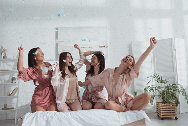 Mujeres felices y emocionadas multiculturales con las manos en el aire en la cama con plumas en la habitación en la despedida de soltera
 - Foto, Imagen