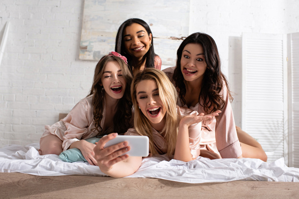 Ευτυχισμένες και ενθουσιασμένες πολυεθνικές γυναίκες που βγάζουν selfie στο κρεβάτι στο μπάτσελορ πάρτι - Φωτογραφία, εικόνα