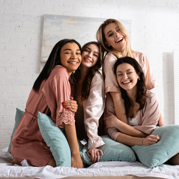 Glückliche multiethnische Freunde blicken in die Kamera, umarmen sich und lächeln zusammen mit Kissen auf dem Bett bei der Junggesellenabschied - Foto, Bild