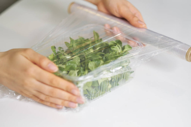Frau verwendet Lebensmittelfolie zur Lagerung von Lebensmitteln auf einem weißen Tisch. Rolle transparenter Polyethylen-Lebensmittelfolie für Verpackungsprodukte. - Foto, Bild