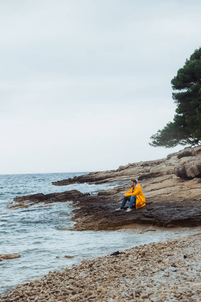 man zittend in gele regenjas op rotsachtig strand kijkend naar stormachtig weer. kopieerruimte. - Foto, afbeelding