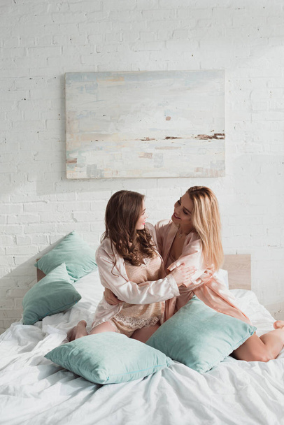 Attraktive Frauen umarmen sich, lächeln und schauen sich bei Junggesellenabschied mit Kissen auf dem Bett an - Foto, Bild