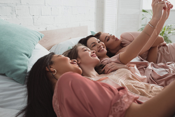 Selektiver Fokus aufgeregter und glücklicher multikultureller Frauen, die ein Selfie machen, während sie sich auf der Junggesellenparty im Bett ausruhen - Foto, Bild