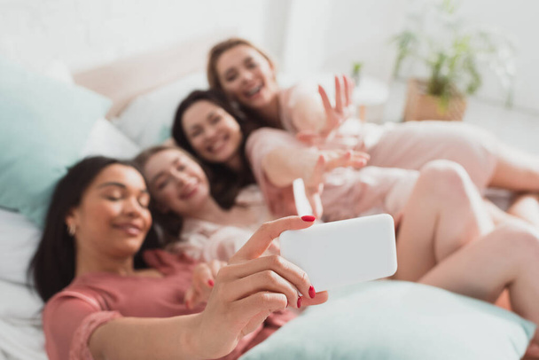 Enfoque selectivo de la chica afroamericana tomando selfie con amigos mientras descansa en la cama en la despedida de soltera
 - Foto, Imagen
