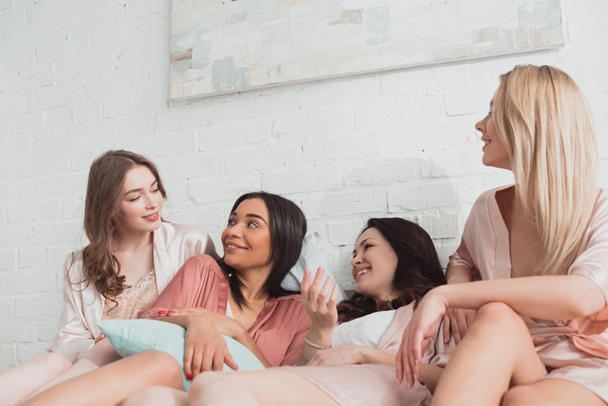Multicultural women looking at each other and smiling on bed at bachelorette party - Foto, immagini