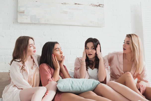 Multicultural women shocked and surprised talking and looking at each other with pillow on bed at bachelorette party - Photo, Image