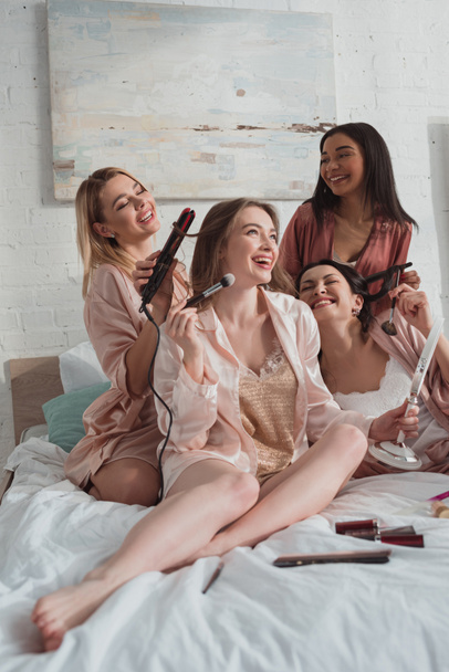 Selektivní zaměření multietnické ženy dávat make-up a dělat účesy na rozlučce se svobodou na posteli - Fotografie, Obrázek