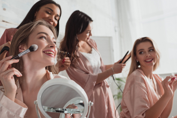 Selektivní zaměření multietnické ženy dávat make-up s kosmetickým štětcem a dělat účesy na pokoji na rozlučce se svobodou - Fotografie, Obrázek