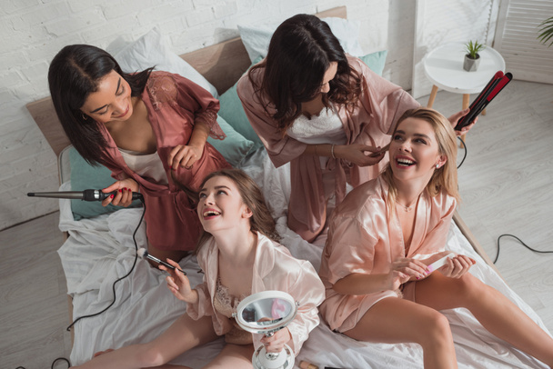 Vue en angle élevé de femmes multiethniques heureux ensemble en utilisant des cosmétiques et en faisant des coiffures sur le lit
 - Photo, image