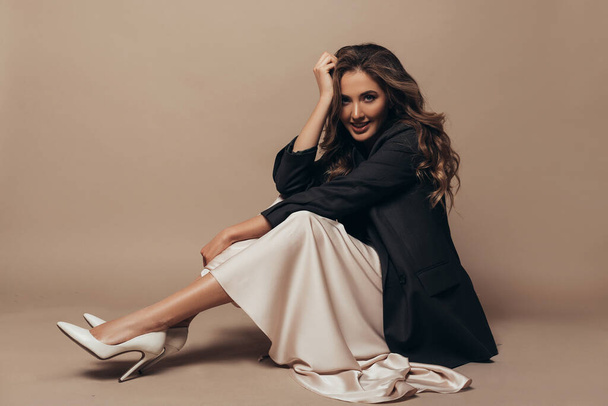 Cheerful model sitting on the floor in studio on the brown background. She wearing modern oversize black jacket and creamy long dress, high heel shoes on her feet. Curly hairstyle and makeup - Photo, Image