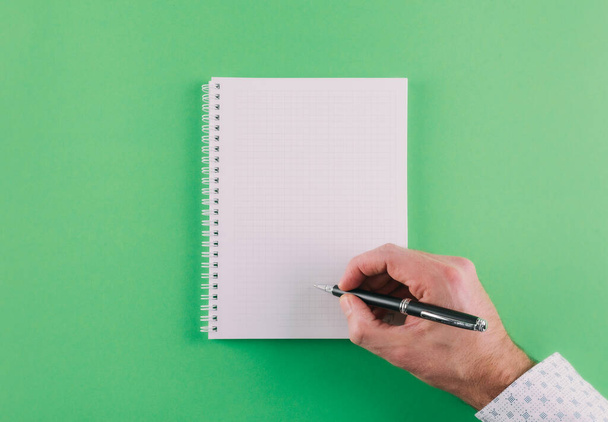 top view of hand holding ballpoint pen against spiral notepad on green background - 写真・画像