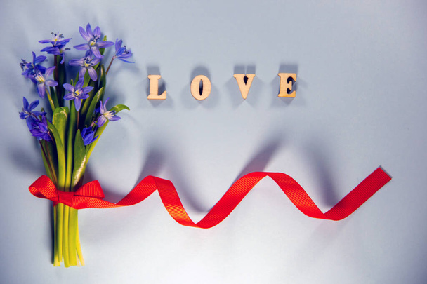 青い背景に赤いリボンでブルーベルの花束新鮮な花。木製の手紙で作られた言葉の愛。トップビュー、フラットレイアウト、コピースペース. - 写真・画像