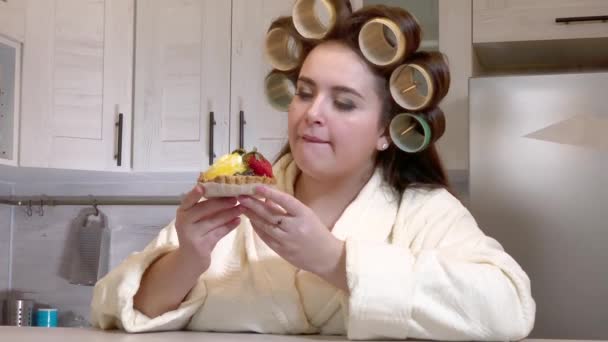 Plus-size girl, dressed in a bathrobe, curlers on her head, looks at the cake with desire, not daring to start eating it - 映像、動画