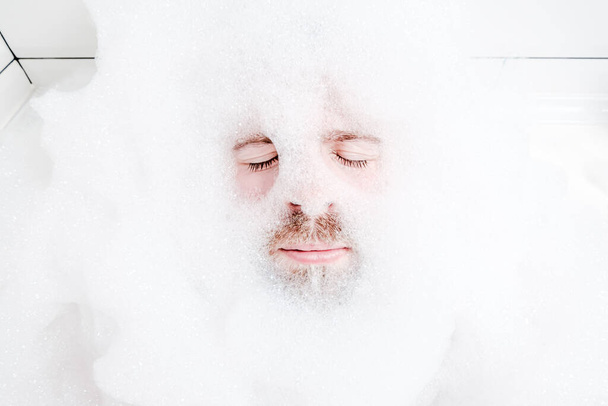 Male face in a lush white foam, only eyes and mouth are visible, close-up. Man relaxes while lying in a bathtub, with soapy water. - Φωτογραφία, εικόνα