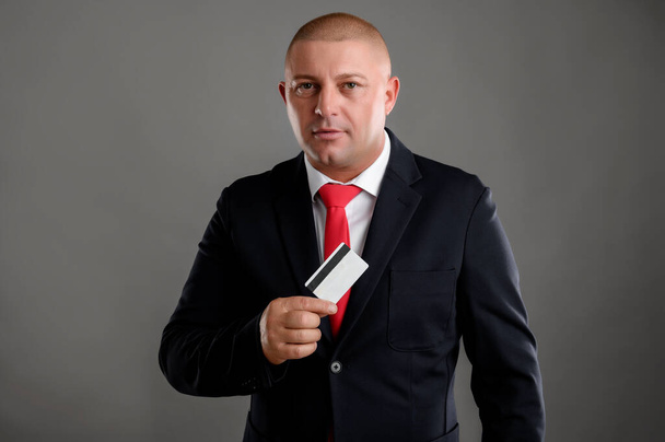 Portrait d'homme d'affaires cool portant élégant costume noir et cravate rouge montre sa carte de crédit isolé sur fond gris
 - Photo, image