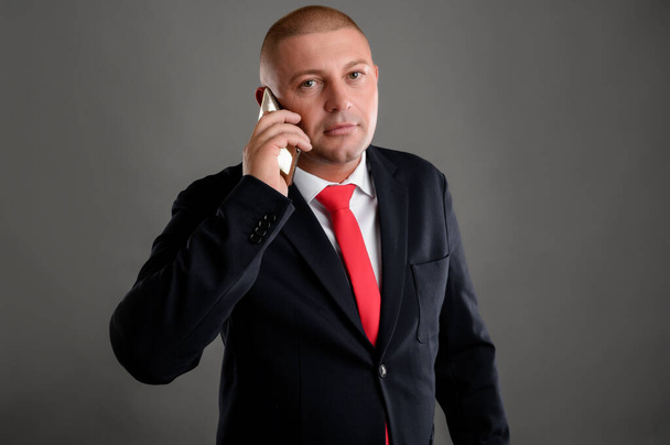 Zarif siyah takım elbiseli havalı iş adamının portresi ve telefonda konuşan kırmızı kravat gri arka planda izole edilmiş. - Fotoğraf, Görsel