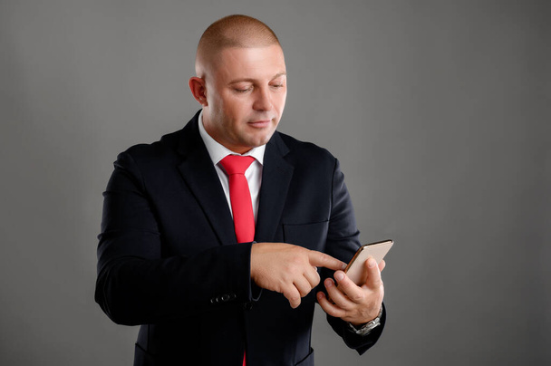 Portrait of cool businessman wearing elegant black suit and red tie using mobile phone, writing text message isolated over gray background - Photo, Image
