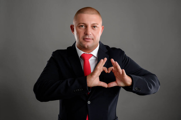 Portrait of cool businessman wearing elegant black suit and red tie making out of hands heart isolated over gray background - Photo, Image