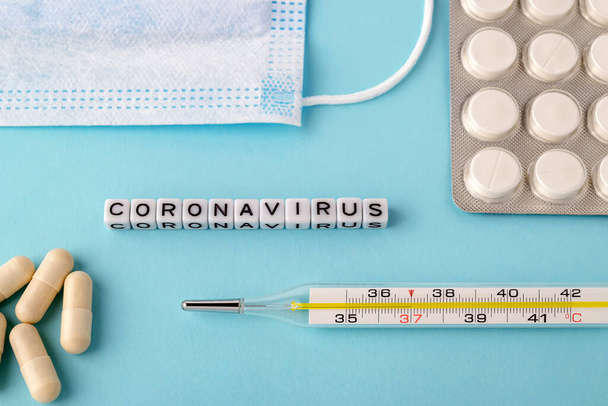 Word Coronavirus, surgical mask, thermometer and pills on a blue background. Medical mask reduces the risk of infection. Airborne disease protection concept. - Фото, изображение