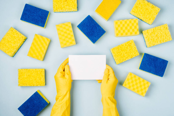 Female hands hold empty card. Blue background with yellow sponges. Cleaning or housekeeping concept background. Copy space. Flat lay, Top view. - Foto, Imagen