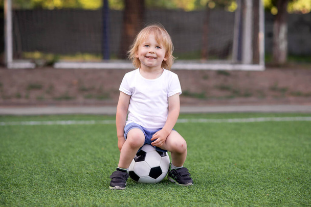 Portrait of blonde toddler kid sitting with broad happy smile on soccer ball at green football field . Nets (goalposts) behind. Little sportsman at playground or soccer lesson in kindergarten or preschool. - Фото, изображение