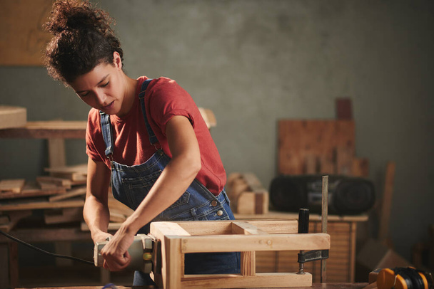 Professional carpenter at work. Young concentrated woman in denim overall smoothing just finished wooden stool with electrical belt sander - Photo, Image