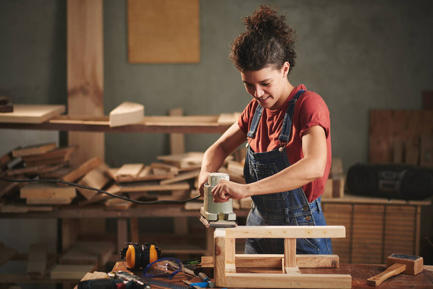 In love with woodworking. Young pretty female carpenter carefully smoothing wooden stool with electrical belt sander and smiling - Photo, Image