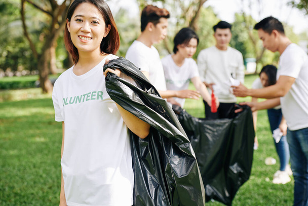 Portrét mladého šťastného dobrovolníka s velkým plastovým pytlem na odpadky pro sběr odpadků v parku - Fotografie, Obrázek