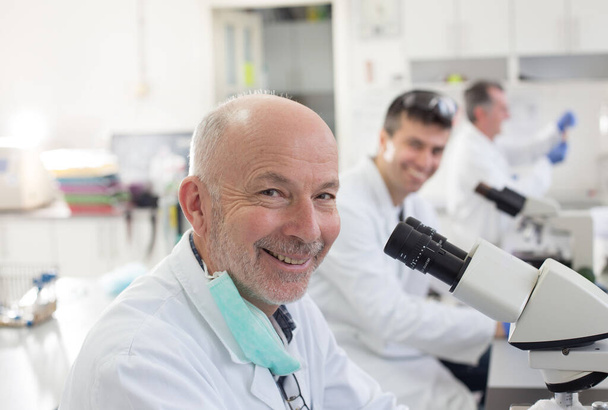 Porträt eines leitenden Biologen, der mit Laborkollegen am Mikroskop arbeitet - Foto, Bild