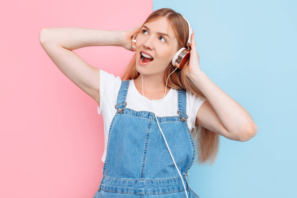 Happy cute beautiful young cheerful pregnant woman enjoys listening to music with headphones while standing on an isolated pink and blue background - Valokuva, kuva