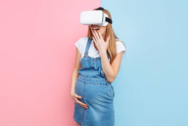 Веселая счастливая беременная женщина получает удовольствие, надевая очки виртуальной реальности, стоя на розовом и синем фоне
 - Фото, изображение