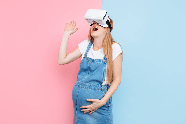 Μια χαρούμενη ευτυχισμένη έγκυος γυναίκα διασκεδάζει φορώντας γυαλιά εικονικής πραγματικότητας, ενώ στέκεται σε ένα ροζ και μπλε φόντο - Φωτογραφία, εικόνα