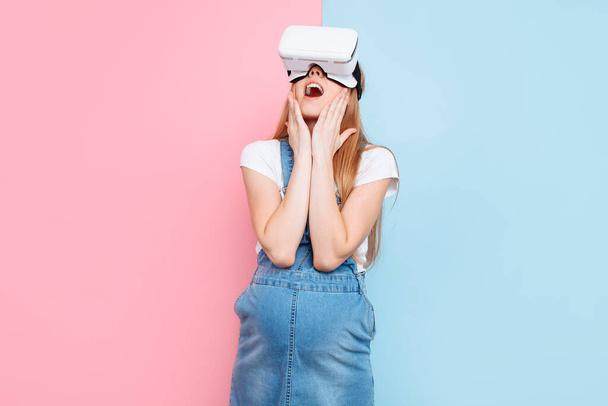 A cheerful happy pregnant woman has fun wearing virtual reality glasses while standing on a pink and blue background - Valokuva, kuva