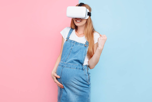 A cheerful happy pregnant woman has fun wearing virtual reality glasses while standing on a pink and blue background - Foto, afbeelding