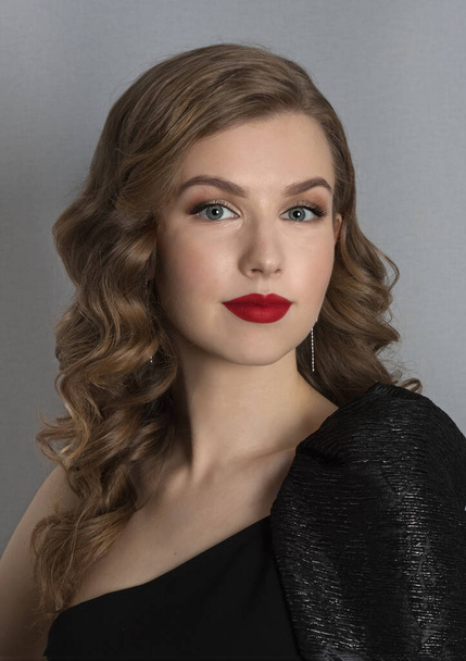 classic portrait of a girl with blond wavy hair in retro style. Young girl in a black dress and with red lipstick on a gray background - Fotoğraf, Görsel